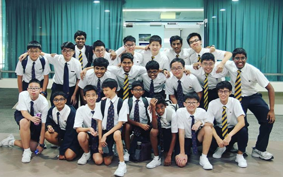 A large group of male students in white school uniforms and striped ties pose for a group photo indoors, arranged in rows with some kneeling in front.